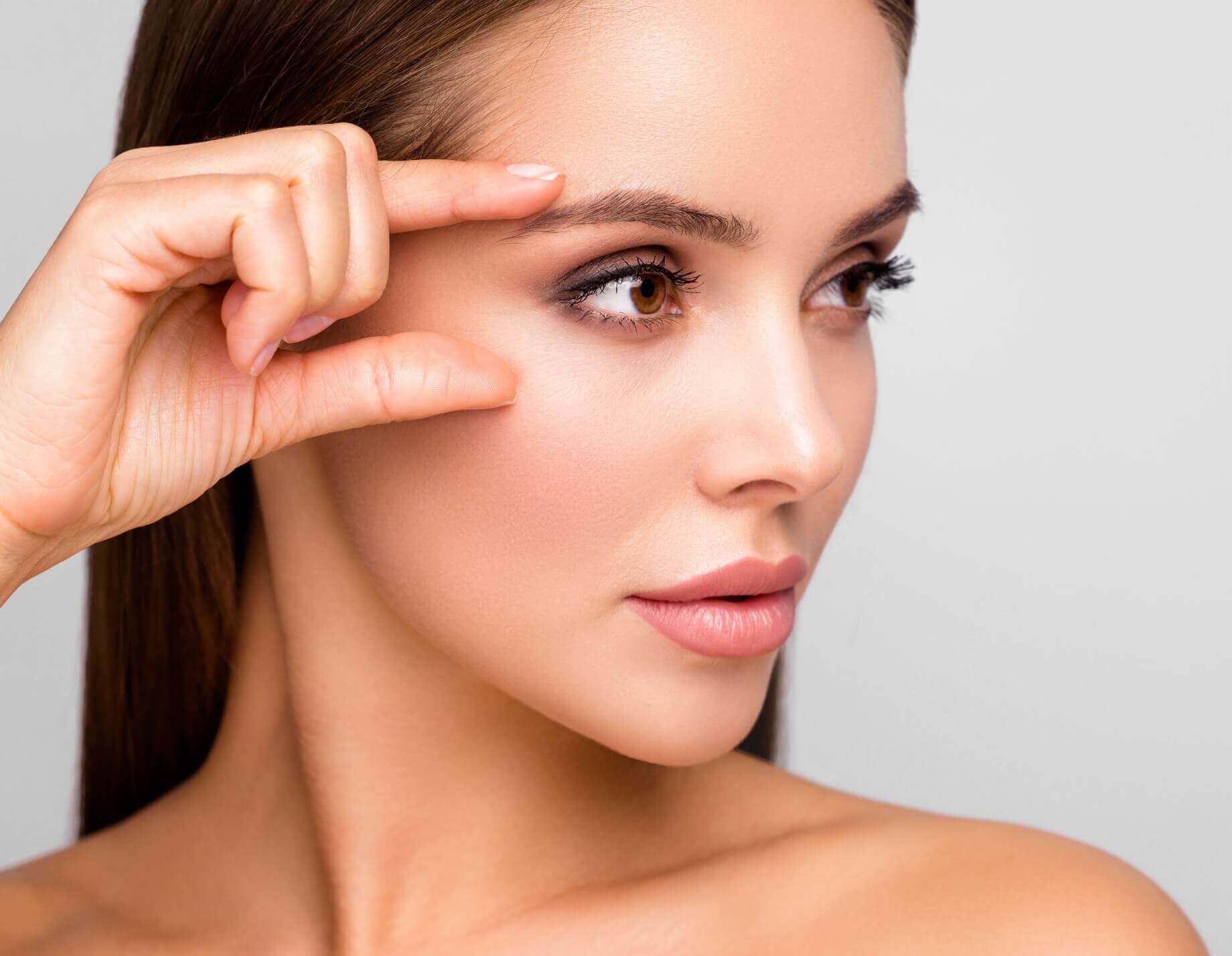 a closeup of a woman with dark brown hair, her right hand framing her eyes, standing against a light grey background