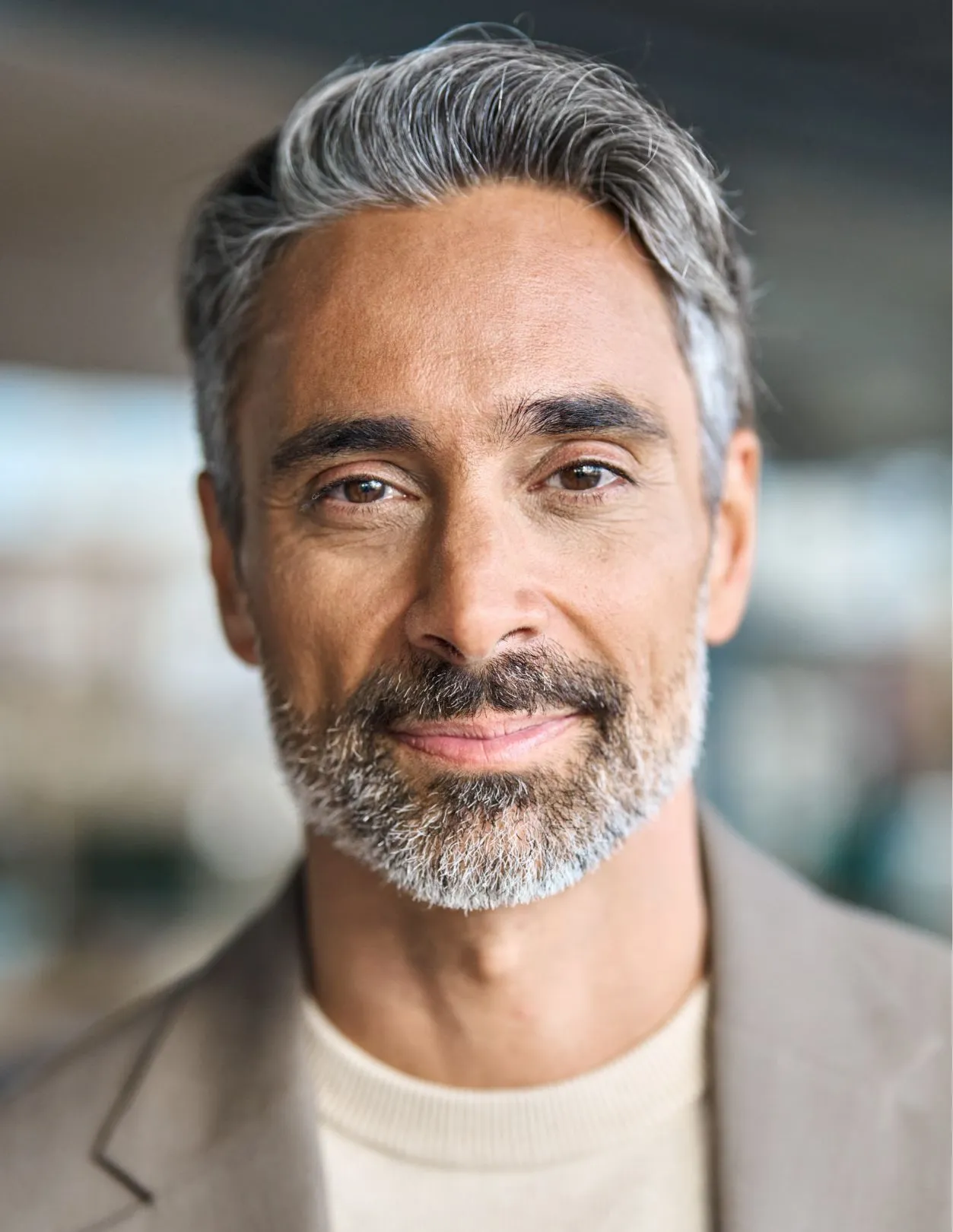 A man with grey hair and a beard, wearing a brown sweater and brown jacket